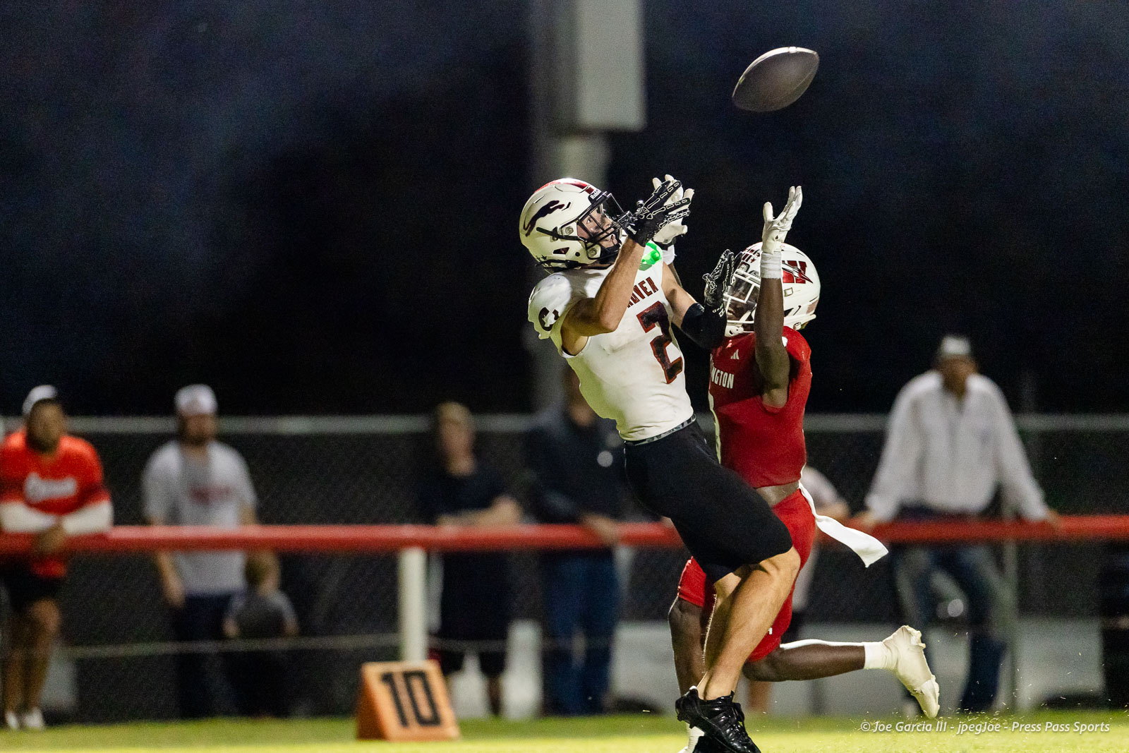 gruver-vs-wellinton-2026-jpegJoe-pps-32 | Press Pass Sports Two football players leap for a catch at night, one in a white jersey (#2) and the other in red, near the sideline with spectators and a '10' marker visible behind them.