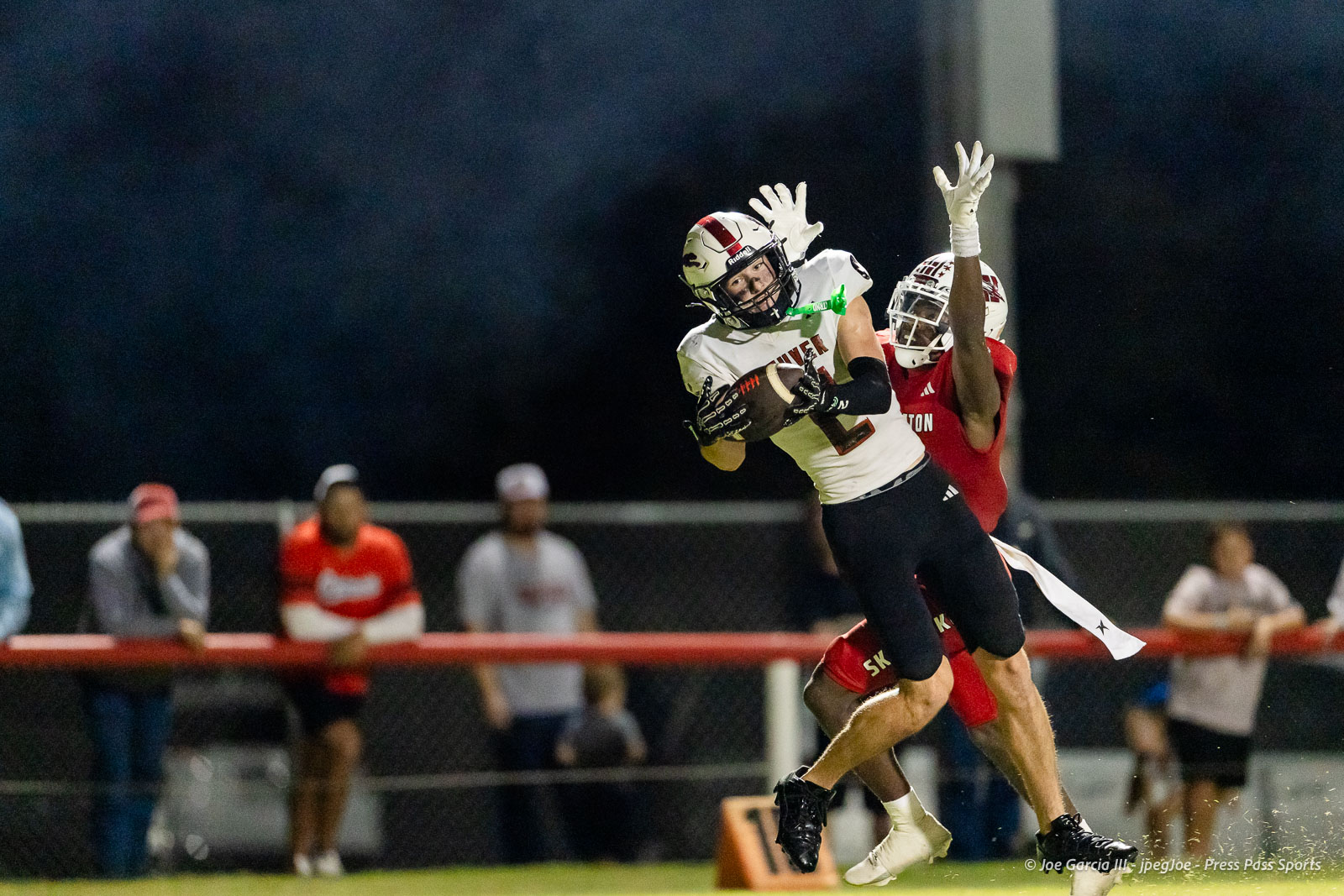 gruver-vs-wellinton-2026-jpegJoe-pps-33 | Press Pass Sports Two football players leap to catch a ball in midair, one in a white jersey and black pants, the other in red, under stadium lights with blurred spectators in the background.