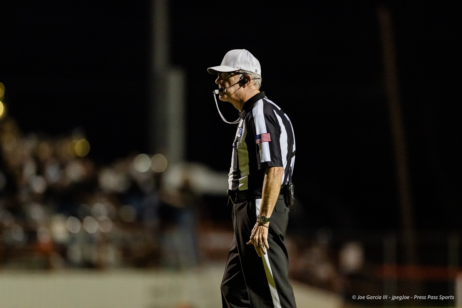 gruver-vs-wellinton-2026-jpegJoe-pps-34 | Press Pass Sports Side view of a football referee in a black-and-white striped uniform, white cap, and headset on a night game with a blurred crowd in the background.
