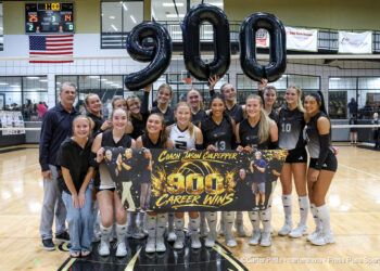 20260926-Dalhart-Bushland-VB-044 | Press Pass Sports Girls' volleyball team poses in a gym holding a banner that says 'Coach Jason Culpepper 900 Career Wins' with large black '900' balloons behind them, celebrating.