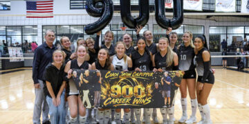 Girls' volleyball team poses in a gym holding a banner that says 'Coach Jason Culpepper 900 Career Wins' with large black '900' balloons behind them, celebrating.