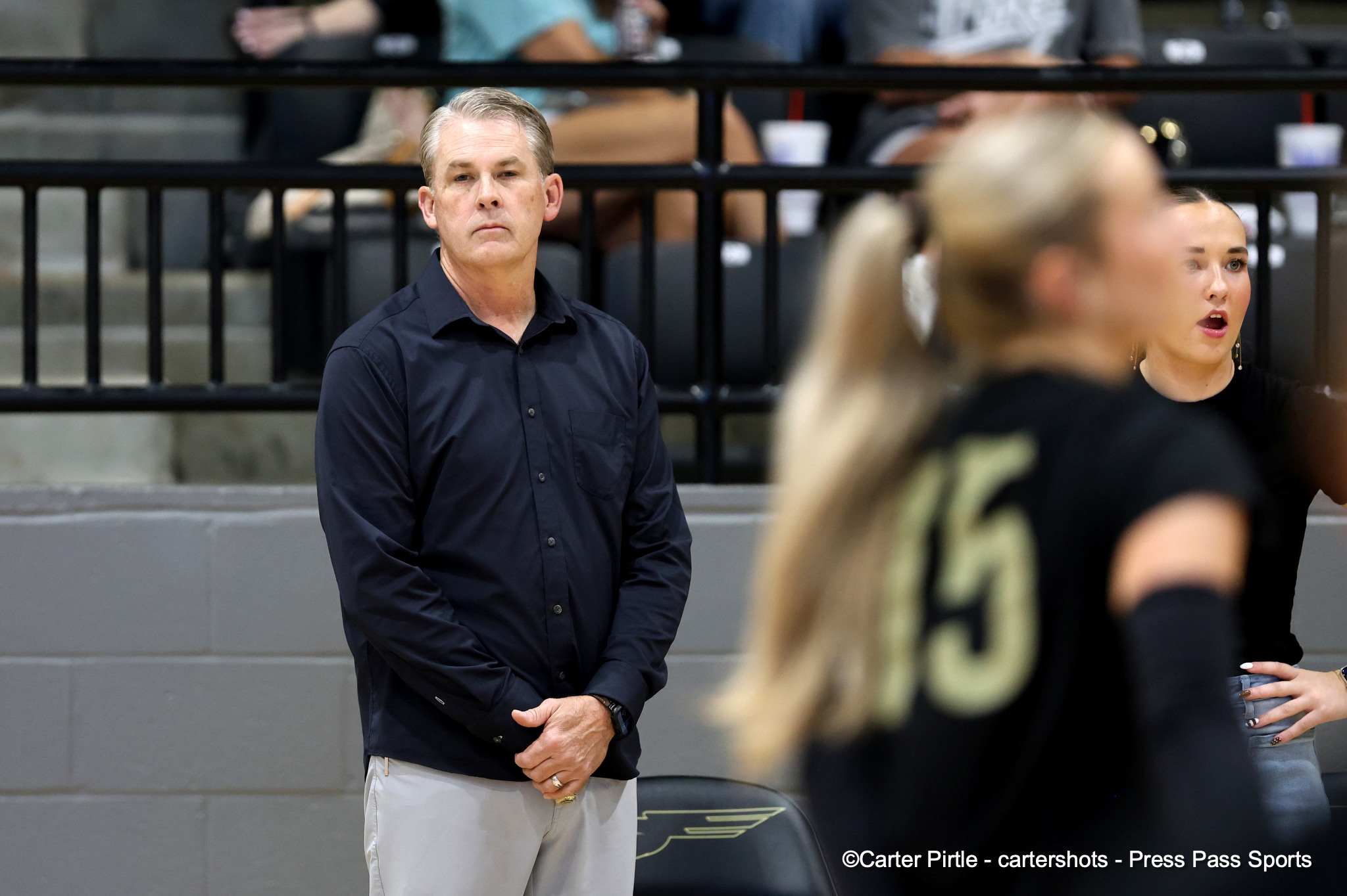 Man in a dark button-down shirt standing with hands clasped in front, inside a gym with blurred spectators in the background.