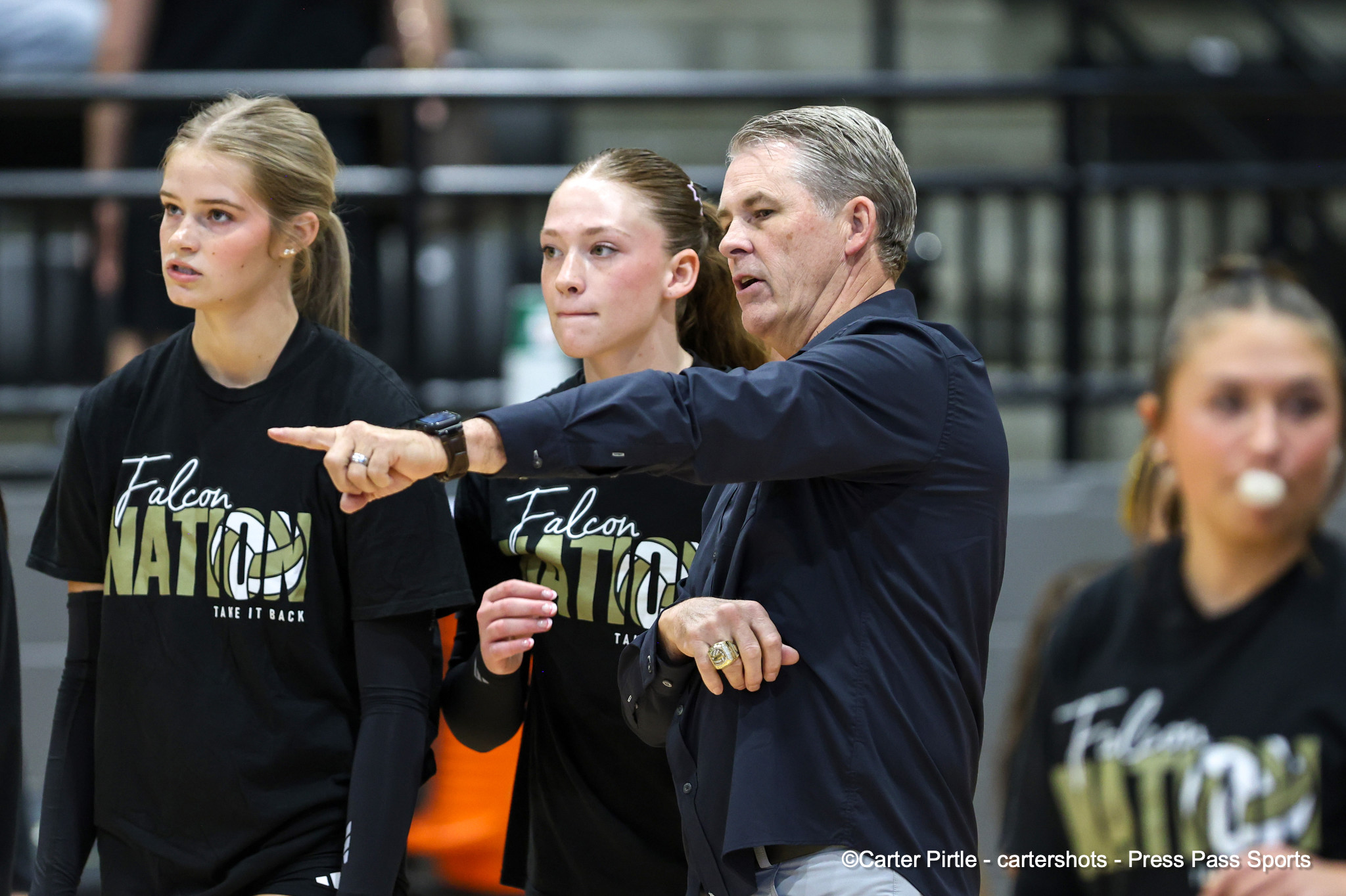 Coach in a dark shirt points as two athletes in Falcon Nation shirts listen in a gym. They stand side by side, focused on something off-camera.