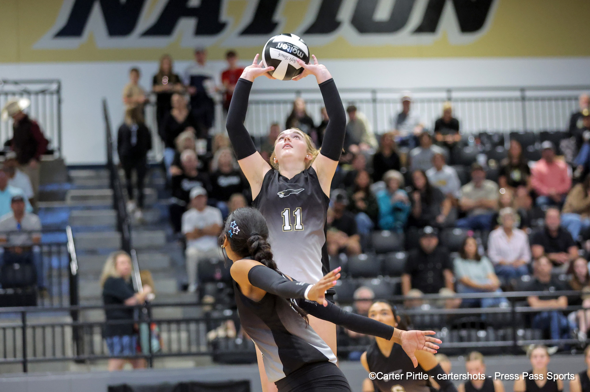 Female volleyball player (number 11) jumps to set the ball overhead during a match; crowd cheers in a gymnasium background.