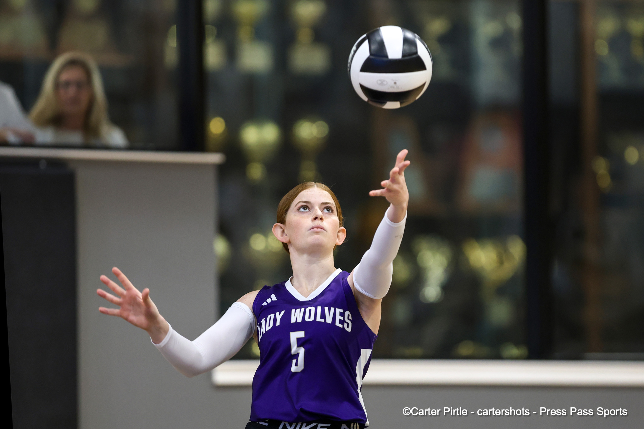 Female volleyball player in a purple jersey (#5) reaches up to contact a volleyball midair, with a blurred gym background.
