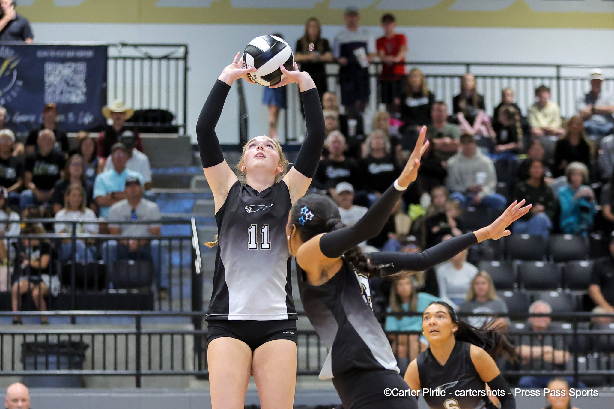 Female volleyball player in a black-and-white uniform raises a ball overhead during a match, crowd in bleachers behind her.