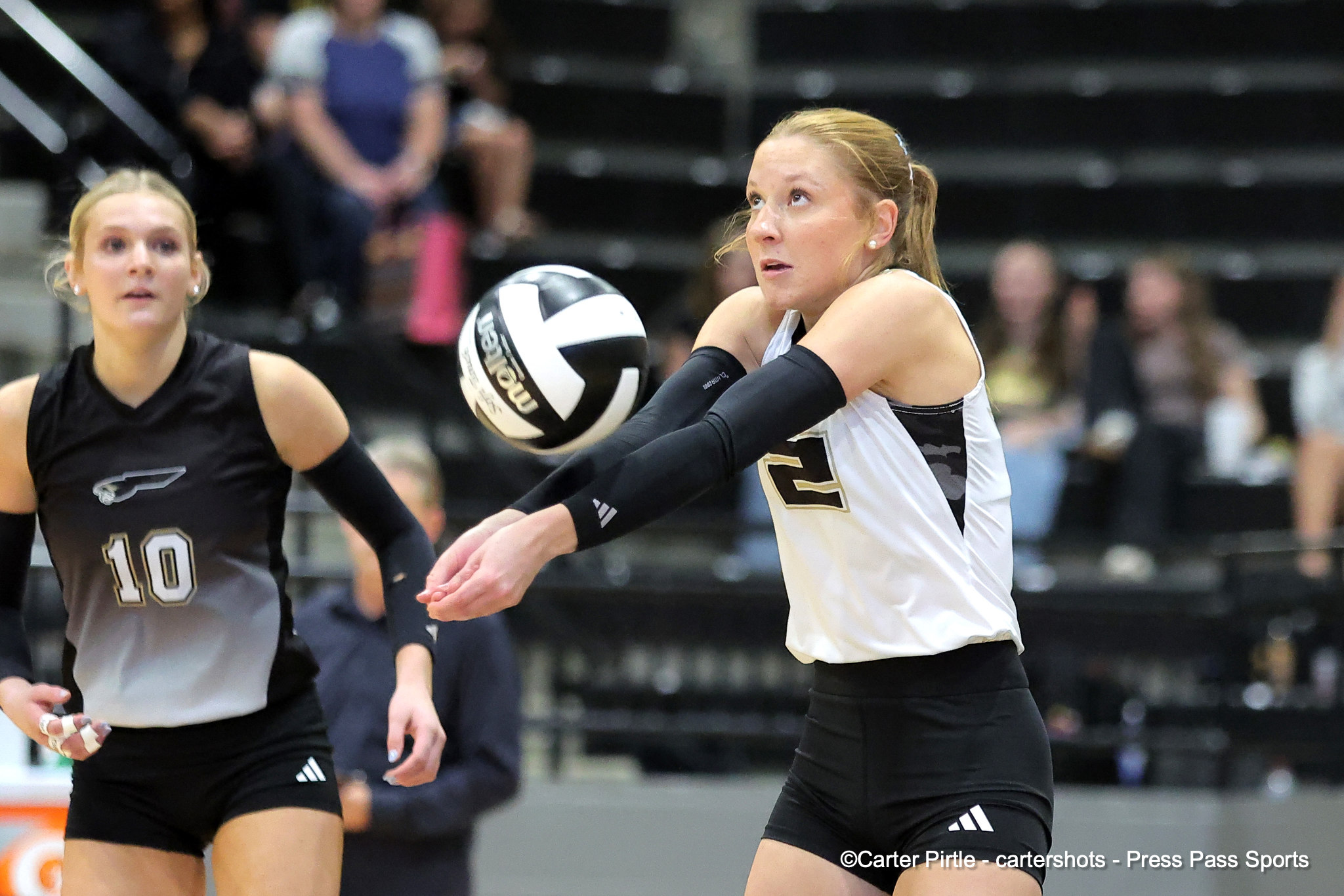 Two female volleyball players chase the ball; the player in white reaches toward a white‑and‑black ball as a teammate in black watches.