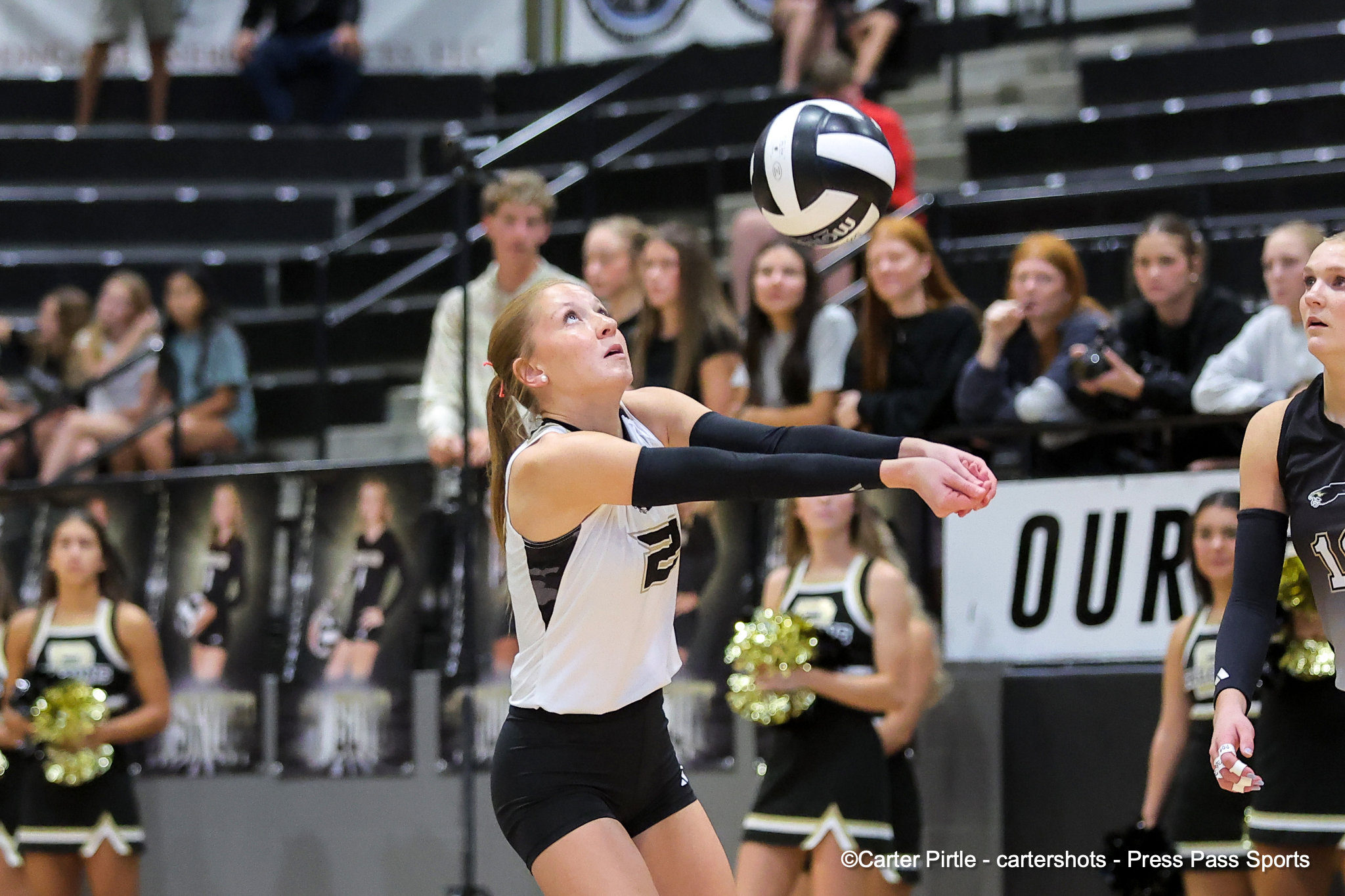 Female volleyball player in a white jersey dives forward to reach the ball as spectators watch nearby.