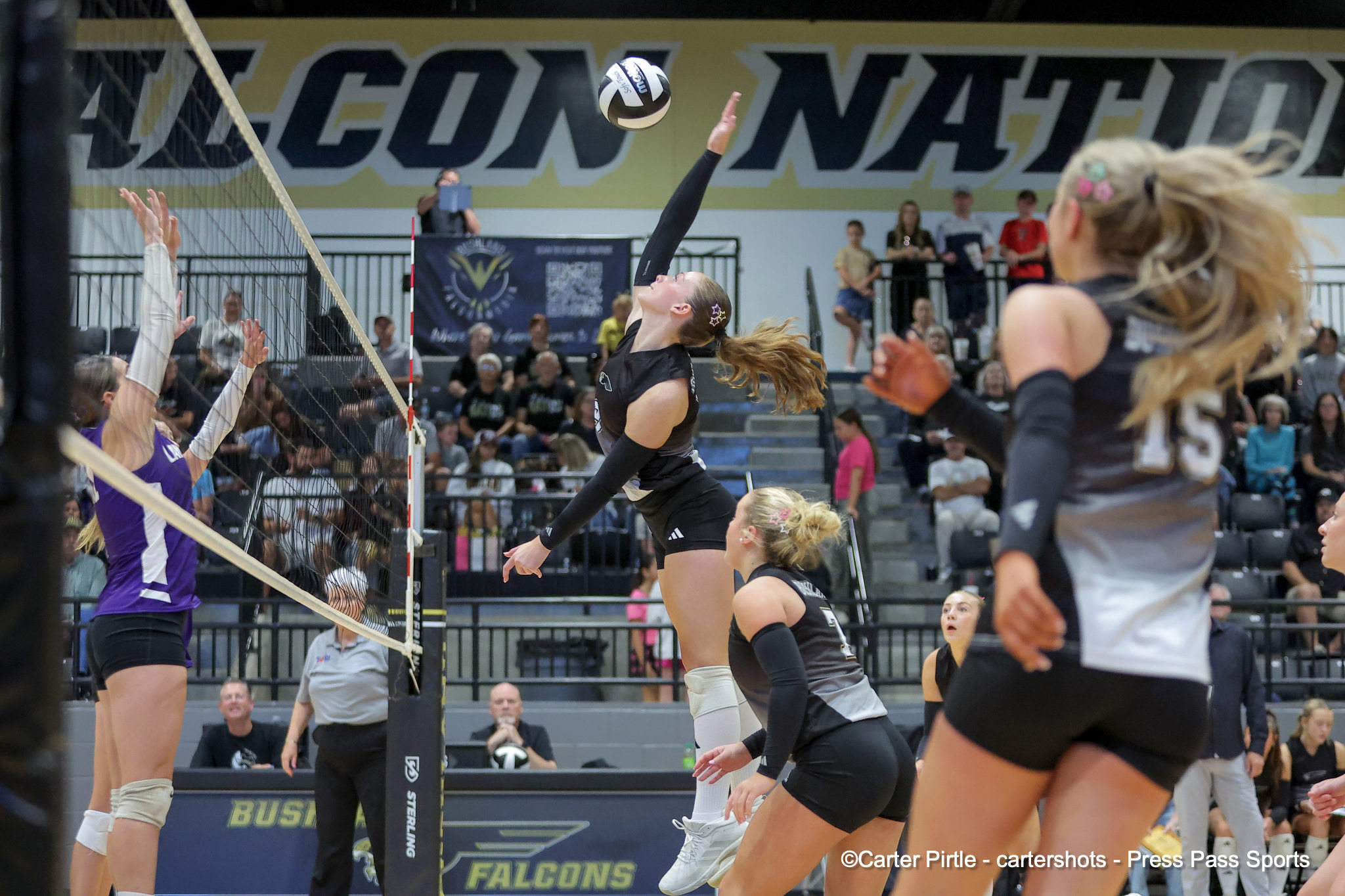 Female volleyball players leap at the net while a teammate sets or spikes the ball during a match in a crowded gymnasium backdrop.