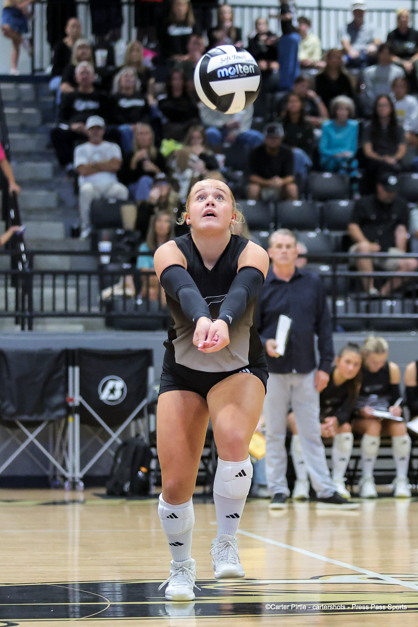Female volleyball player in a black uniform extends forearms to bump a ball overhead, with spectators in the stands watching the play.