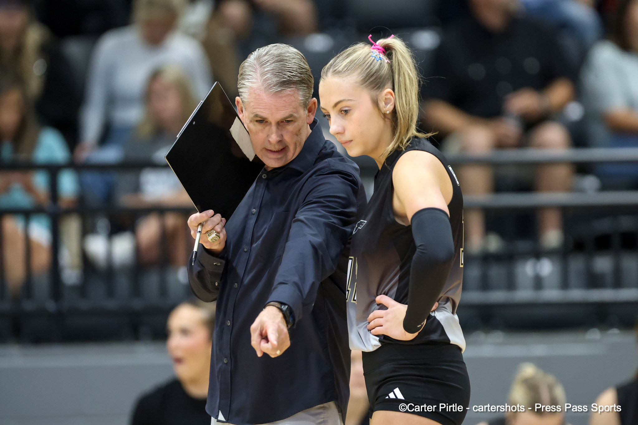 Coach holding a clipboard beside a female athlete in black athletic wear during a game, both focused on something off-camera in a gym arena.