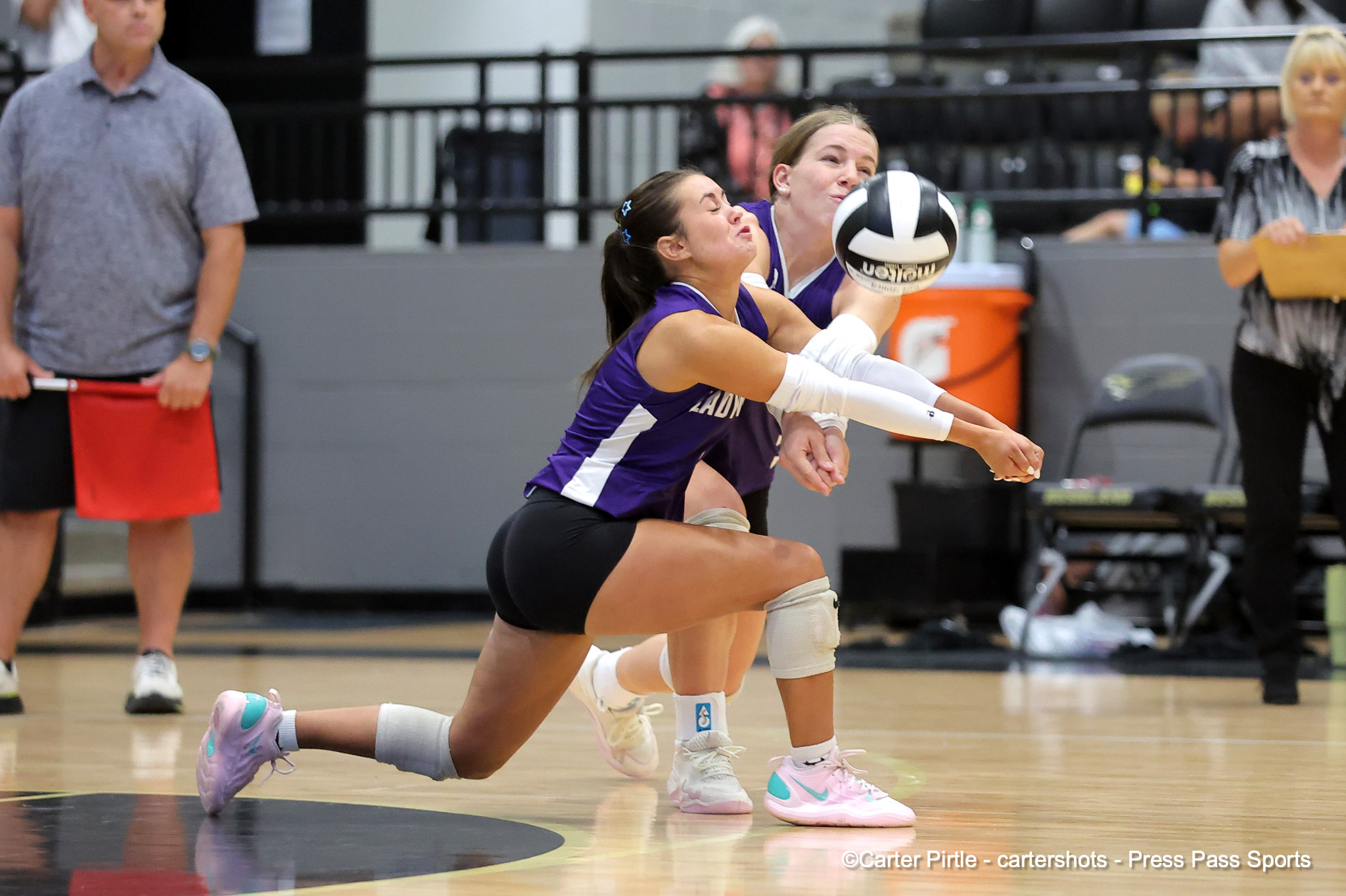 Two female volleyball players in purple uniforms dive for the ball on a gym court, reaching out as a teammate watches nearby.