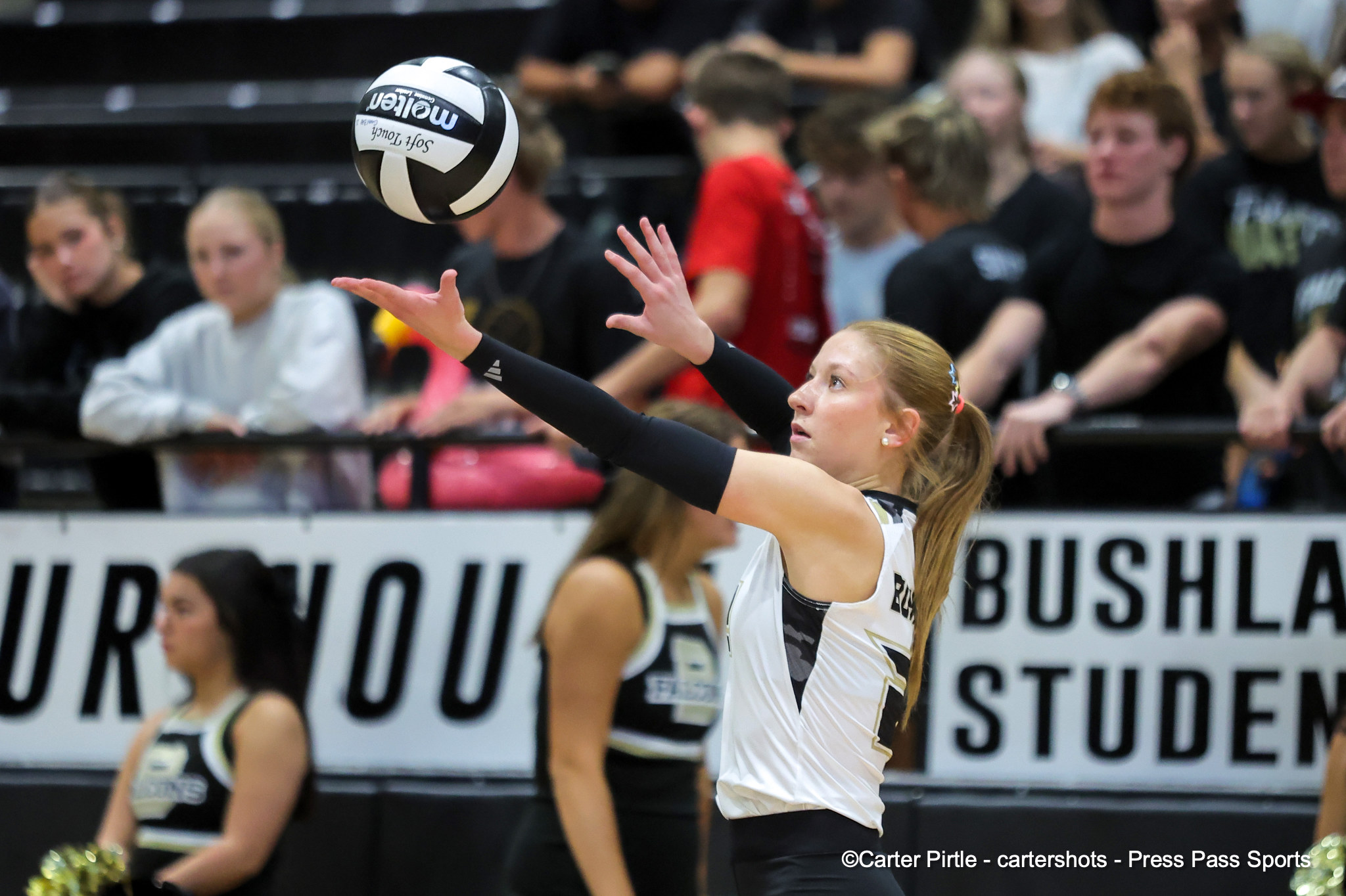 Female volleyball player in white and black uniform extends arms to hit a ball in midair, with a crowd in the stands in the background.