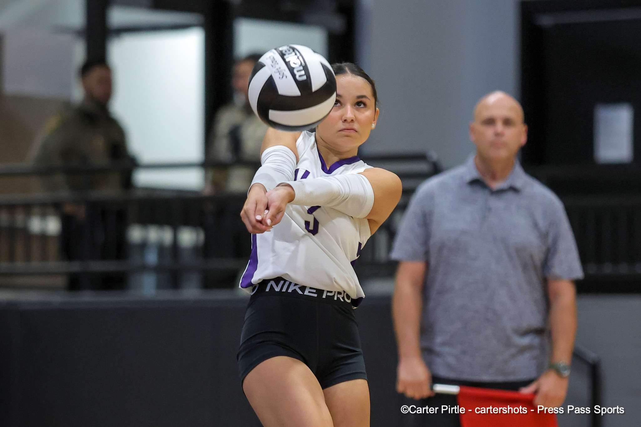 Female volleyball player in a white and purple uniform prepares to spike a ball indoors, focused on the ball with a spectator in a gray shirt in the background.