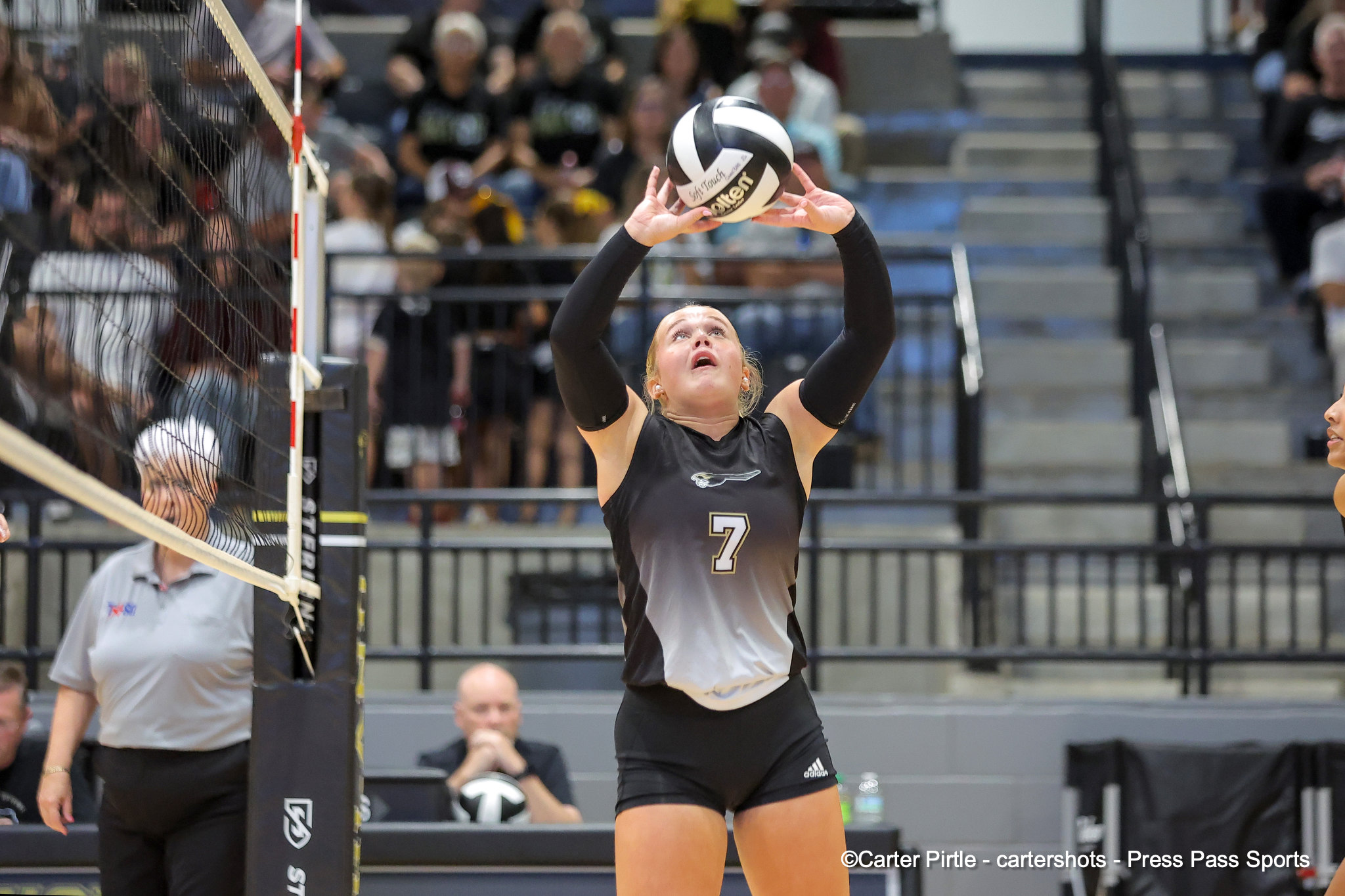 Female volleyball player in black uniform (number 7) reaching up to set a white/gray ball near the net with a crowd watching in the stands behind her.