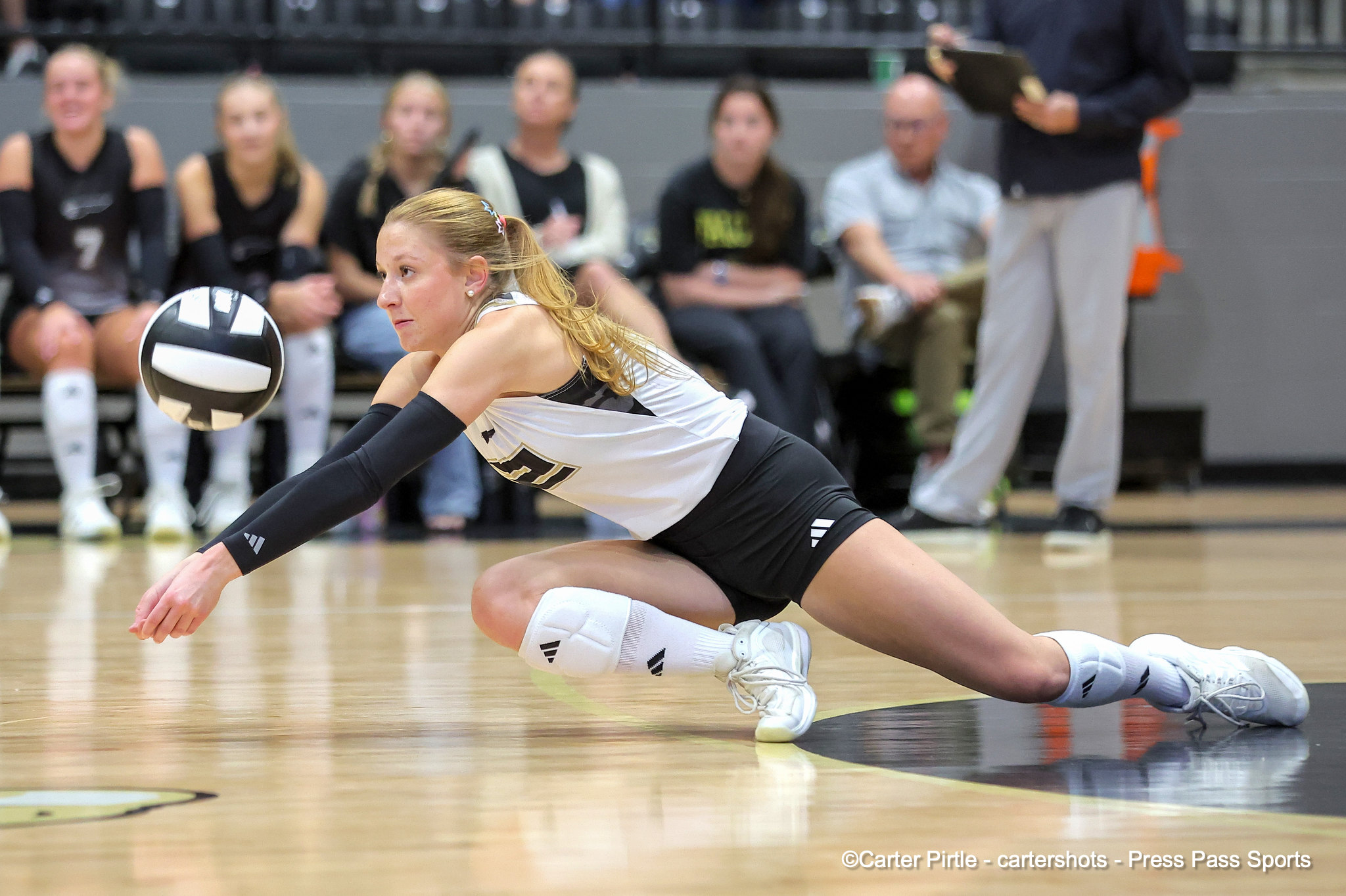 Female volleyball player in a white uniform lunges for the ball on a wooden court, with a blurred bench of spectators in the background.