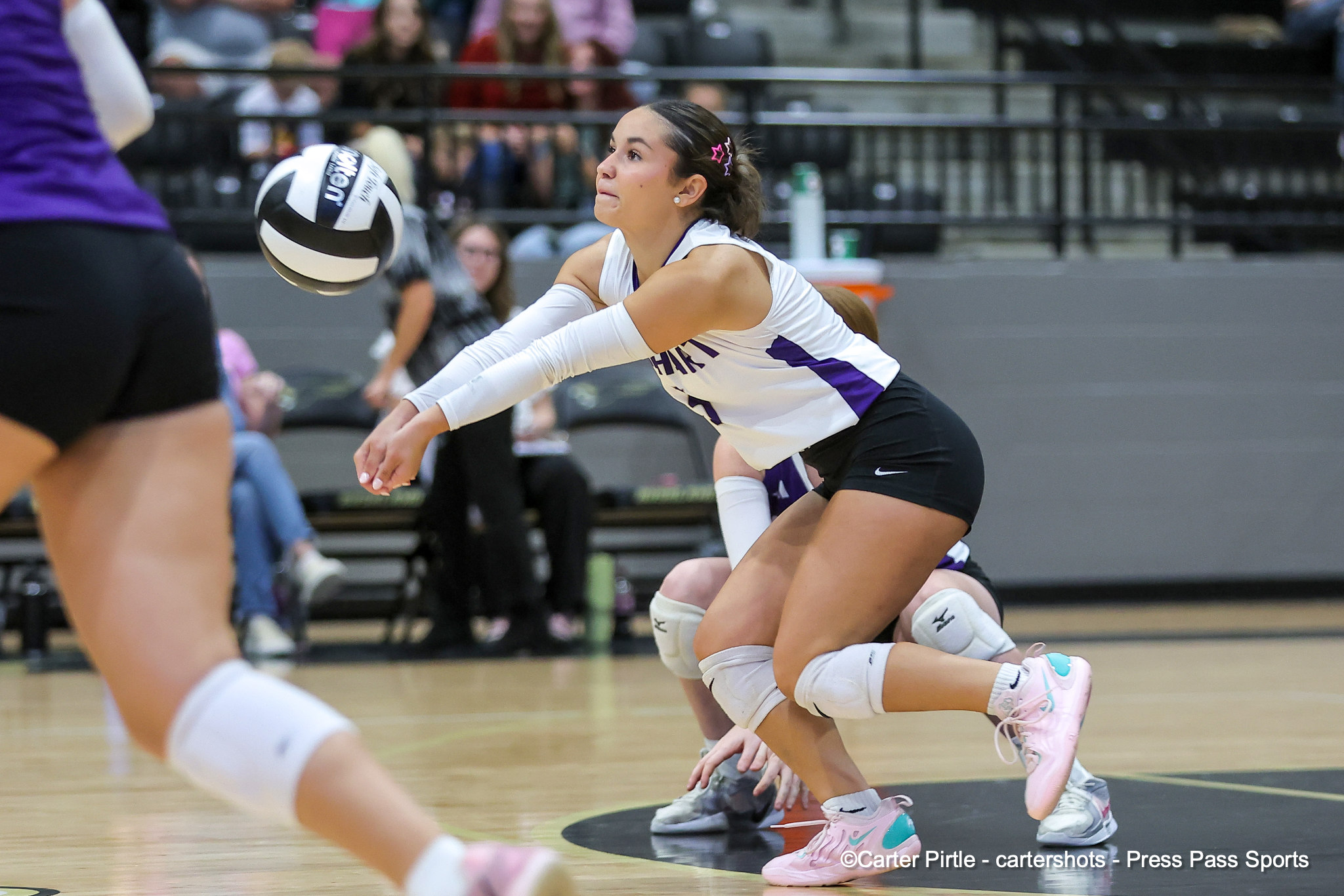 Female volleyball players in white and purple uniforms dive for the ball on a gym court with a volleyball in midair, spectators in bleachers in the background.