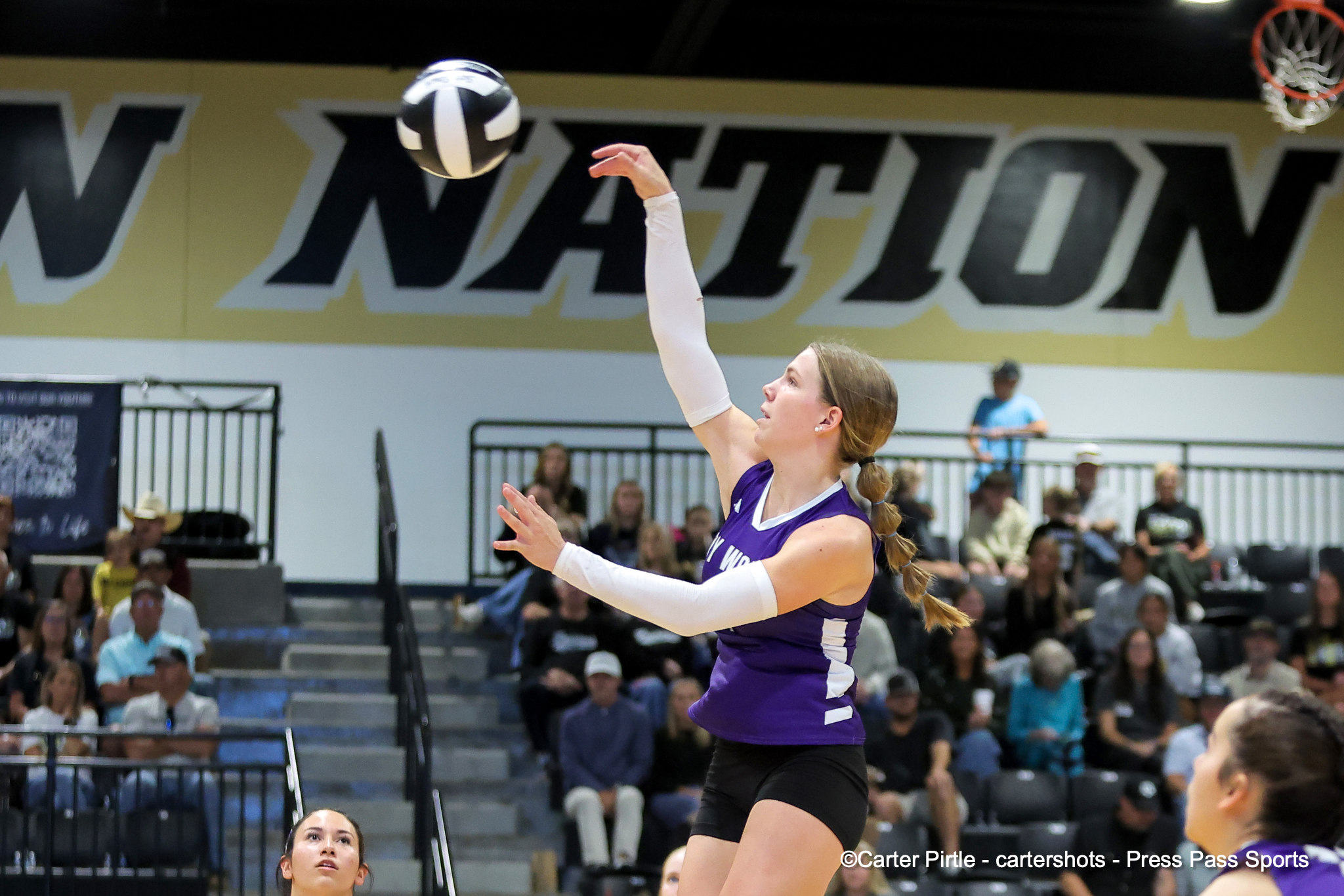 Female volleyball player in a purple uniform reaches up to hit the ball mid-air during a match in a gym filled with spectators.