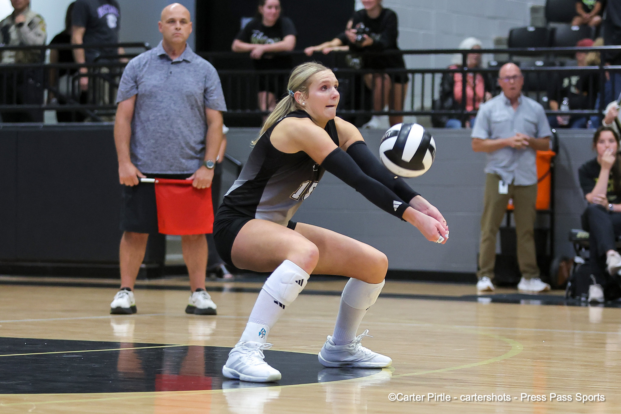 Female volleyball player in a black-gray uniform lunges to receive the ball during a match, gym bleachers in the background.