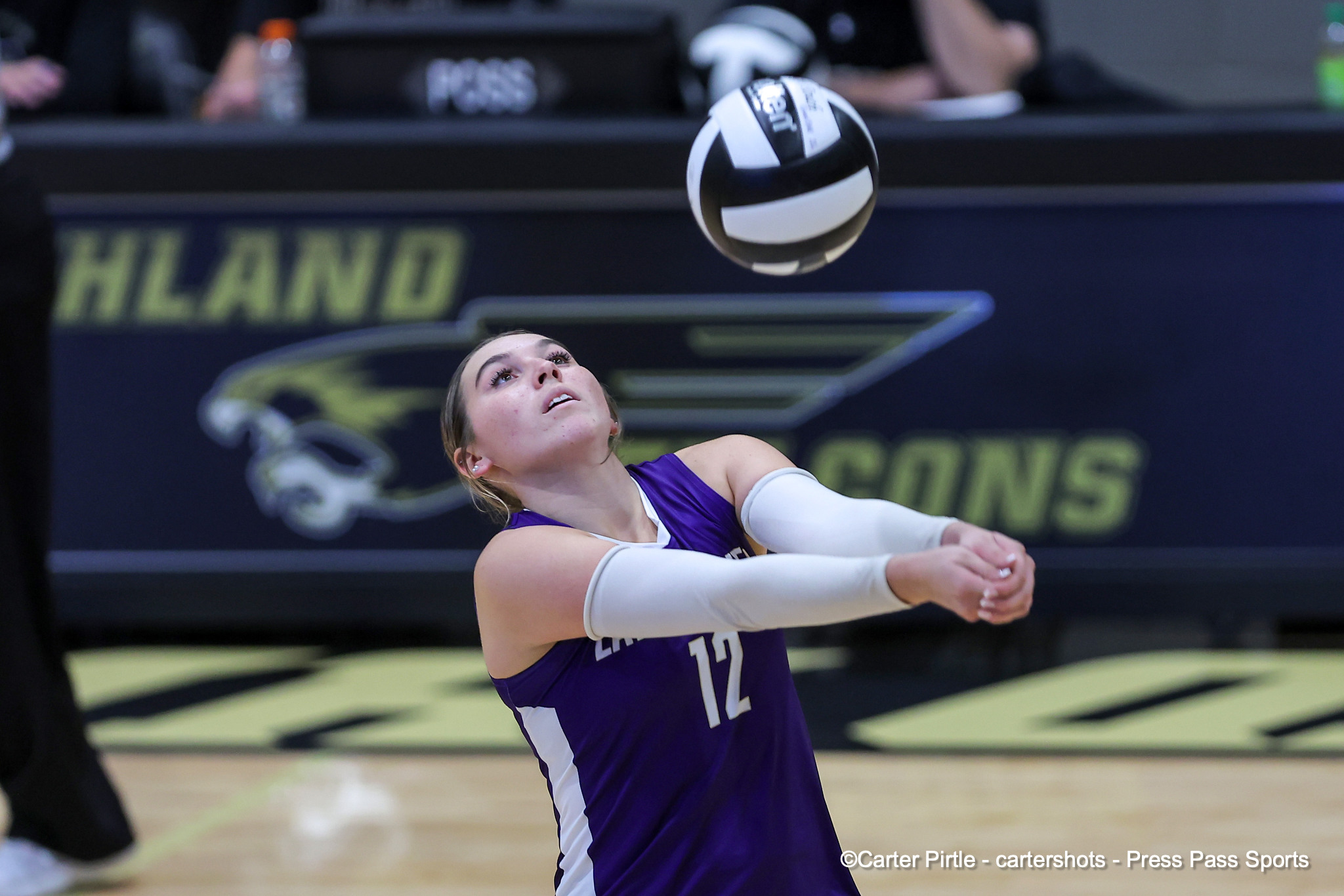 Female volleyball player in a purple jersey extends arms to hit a black-and-white ball mid-air during a match on a court with a banner in the background.