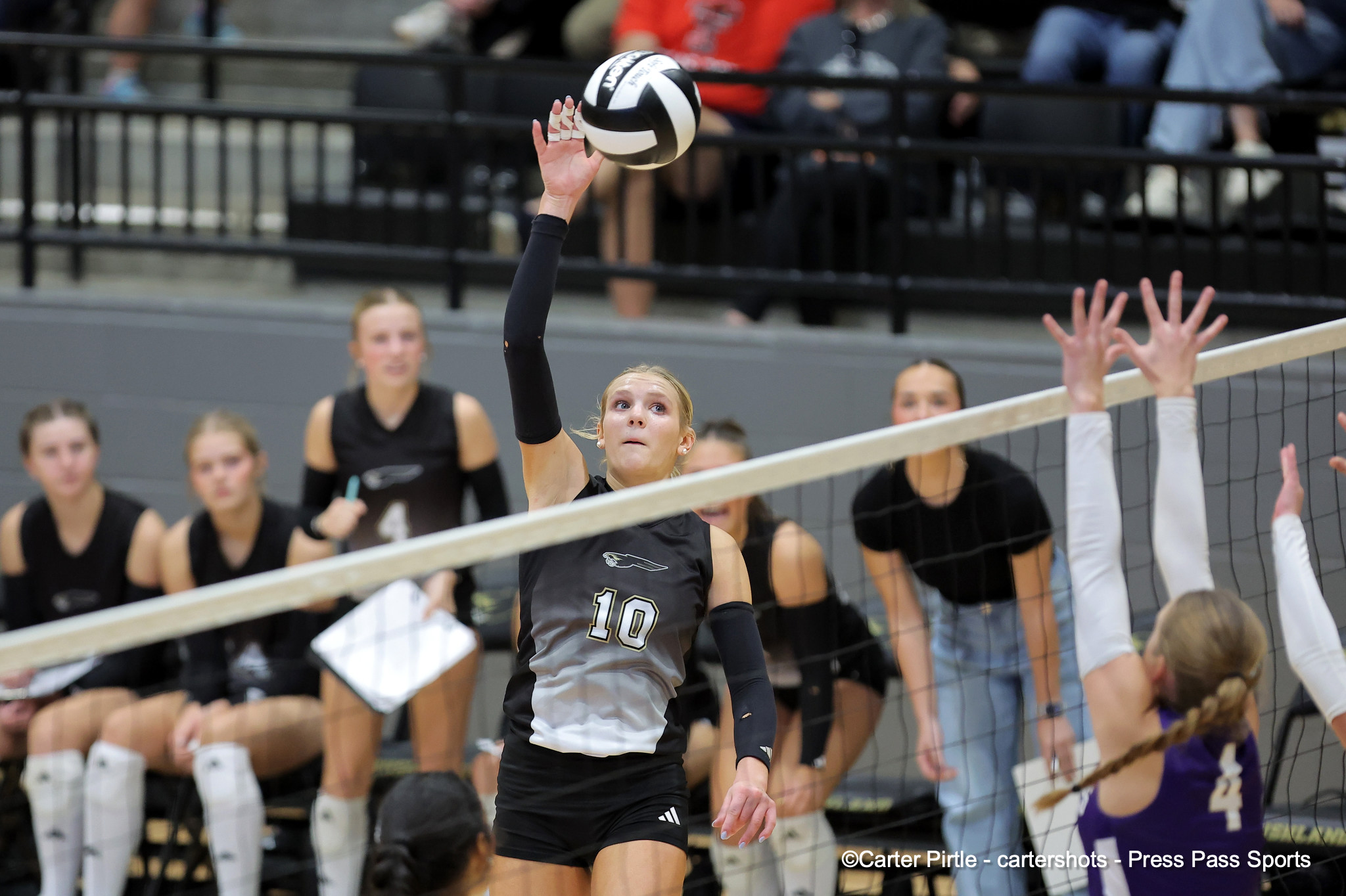 Female volleyball player wearing number 10 spikes the ball over the net during a match, with teammates and spectators in the background.