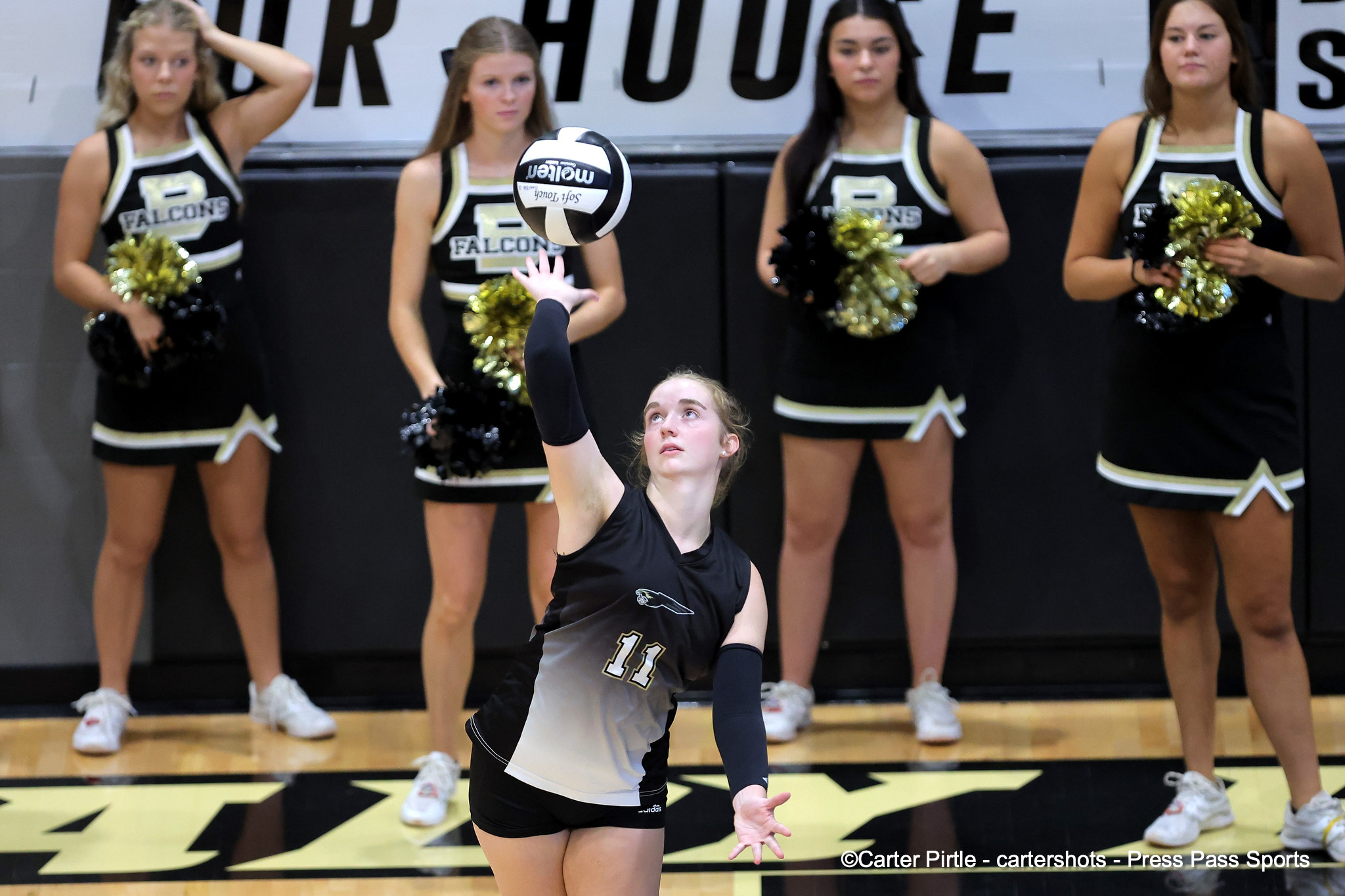 Female volleyball player in a black/gray jersey (#11) serves a ball as four cheerleaders in black uniforms watch from the baseline.
