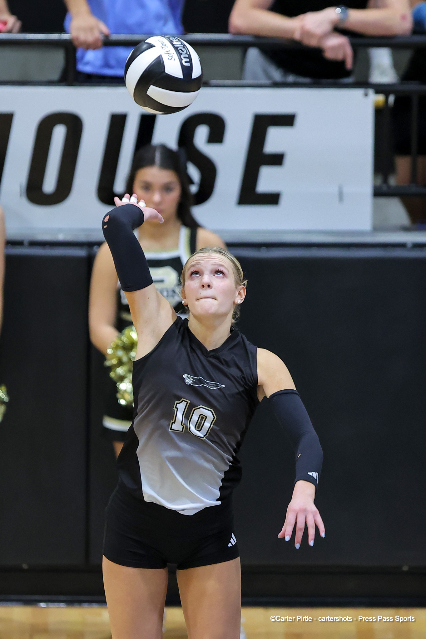 Female volleyball player in black/gray uniform #10 serves the ball during a match, arm raised and ball in midair.