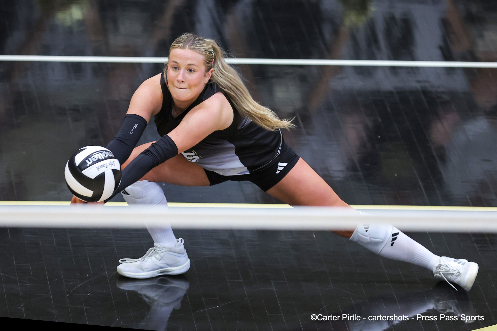 Female volleyball player dives low to reach a white ball on a dark court, extending an arm in a save attempt.