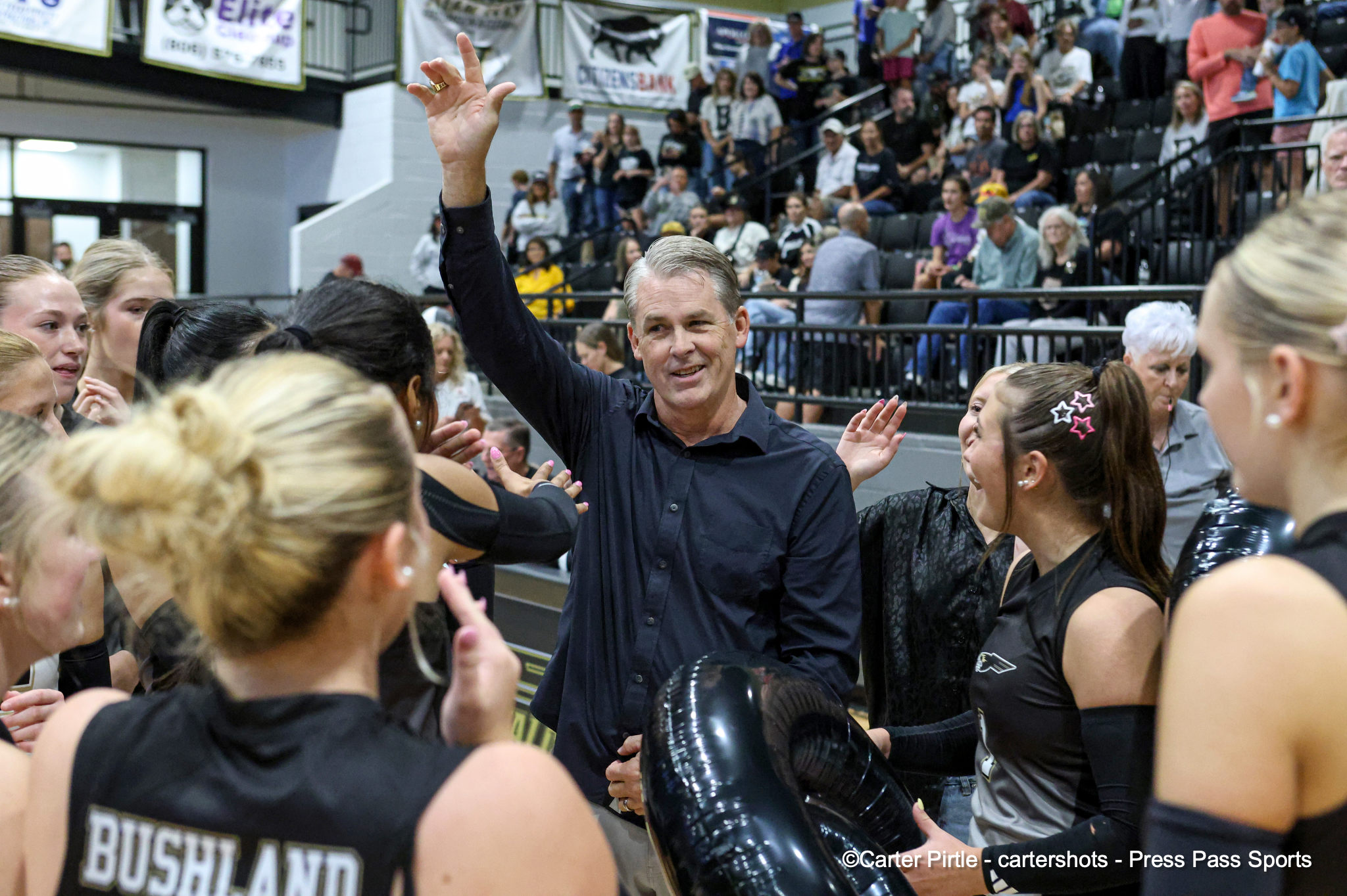 Smiling man in a dark shirt raises his right hand in a wave among a crowd of cheering women at a crowded indoor gym.