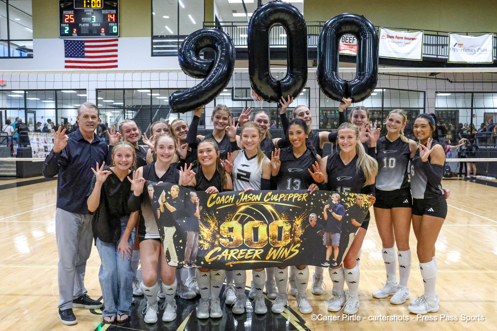 Group of female volleyball players and a coach posing with a banner that reads'Coach Jason Culpepper 900 Career Wins' in a gym, American flag on the wall behind them, and large black number balloons forming 900 above their heads.