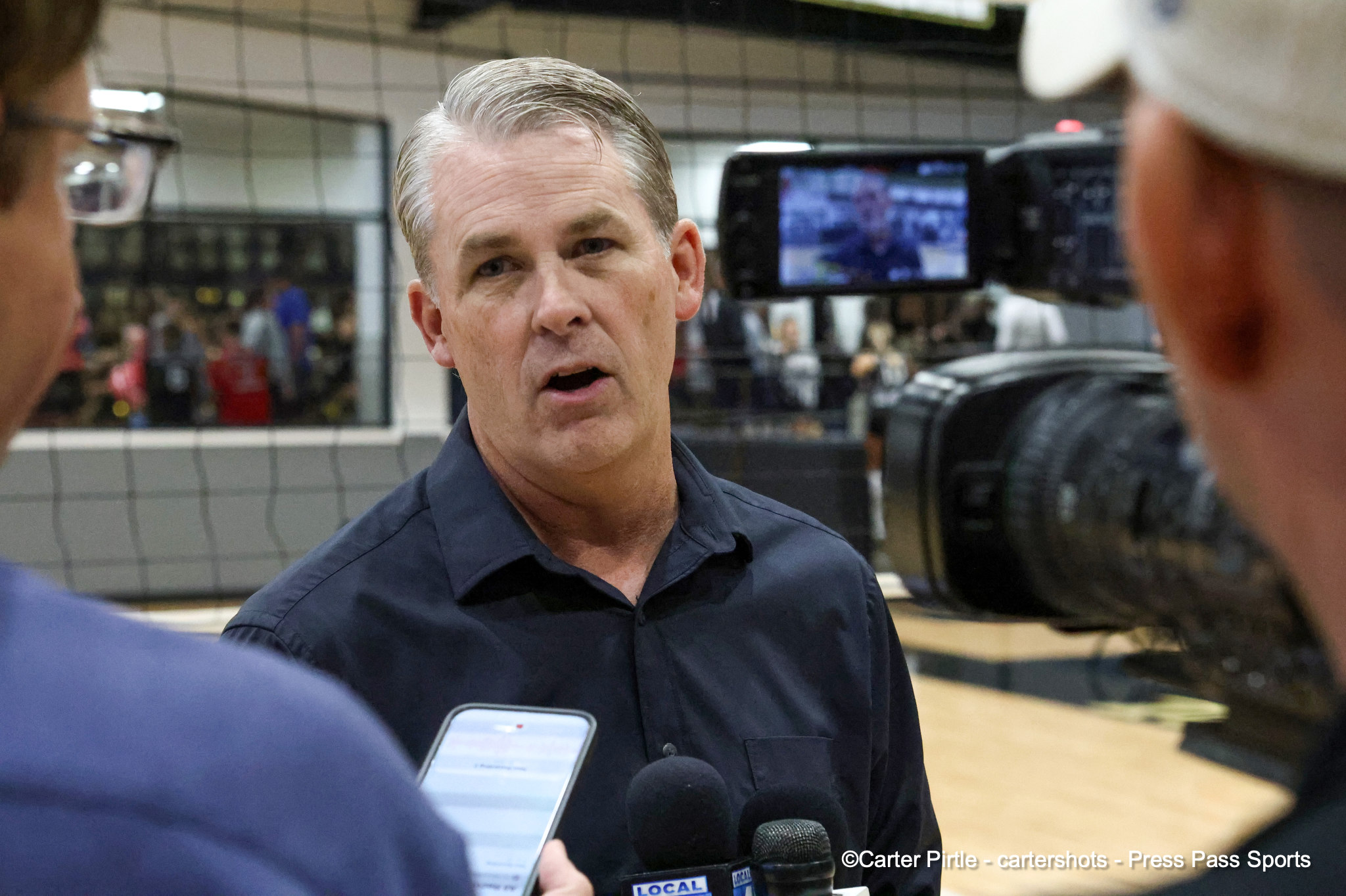 Man in a dark blue shirt answers questions during an on-site interview; microphones near his chest with a camera monitor in the background.”,