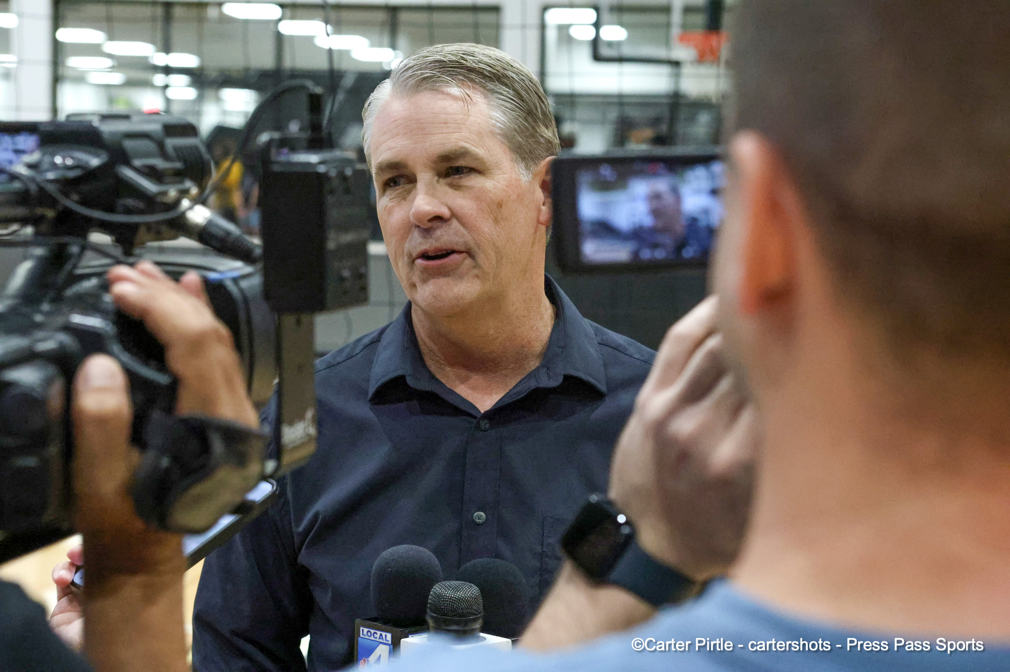 Man in a dark button-down shirt speaks to reporters, several microphones in front amid a newsroom backdrop.