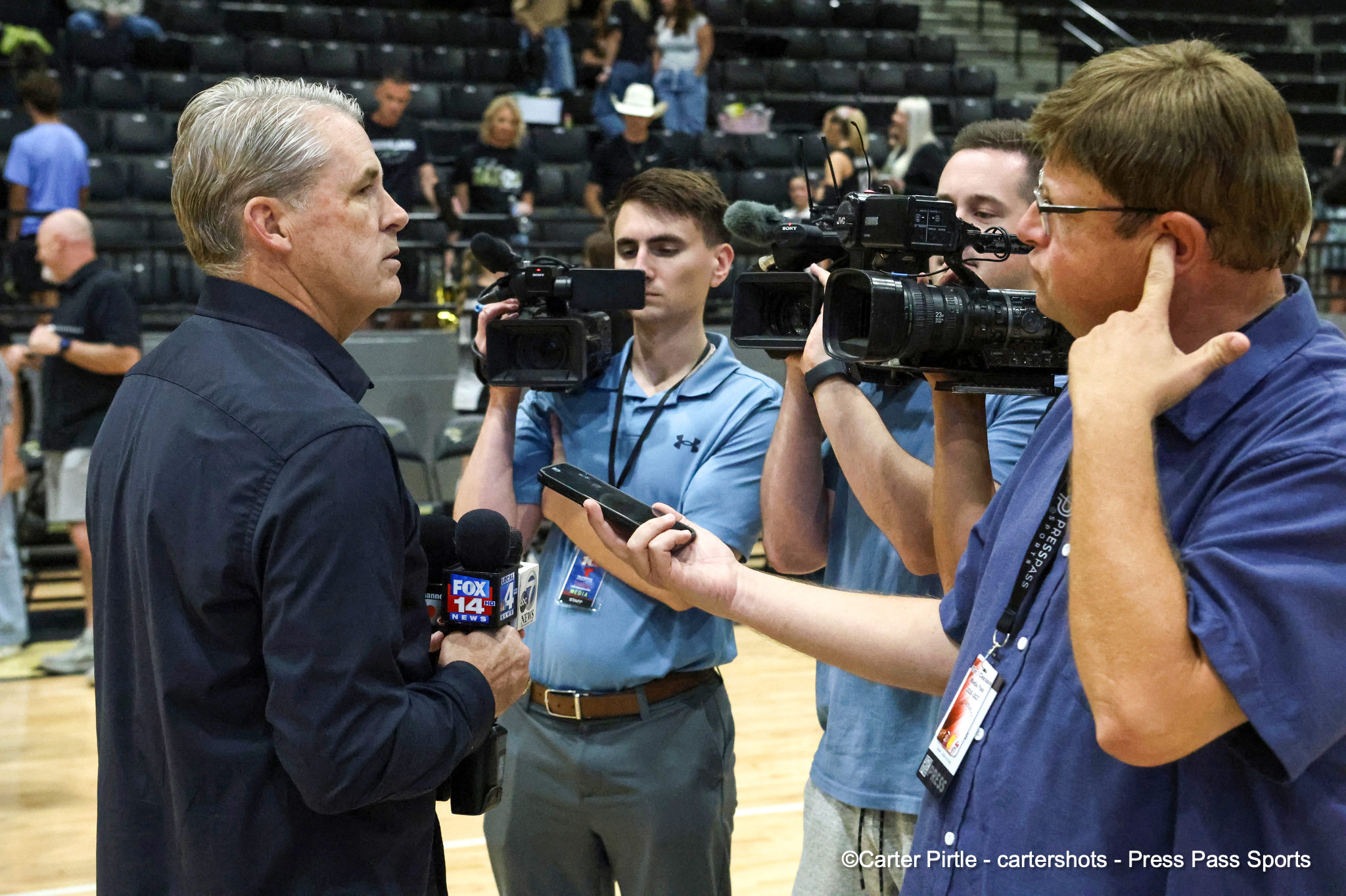 Man in dark shirt being interviewed by reporters holding microphones in a gym arena with cameras pointed at him, FOX 14 News logo visible on mic.