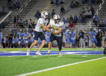 0O3A7075 | Press Pass Sports Two football players in white jerseys run with the ball on a bright green field, crowd watching from the stands behind them.