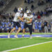 Two football players in white jerseys run with the ball on a bright green field, crowd watching from the stands behind them.