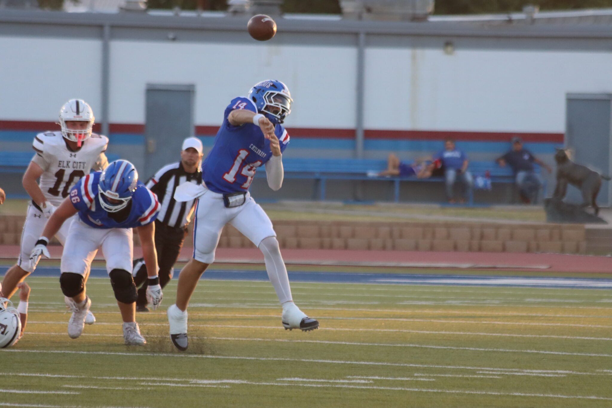 Blue-uniform football player jumps to catch a spiraling ball as defenders watch on a sunny field.