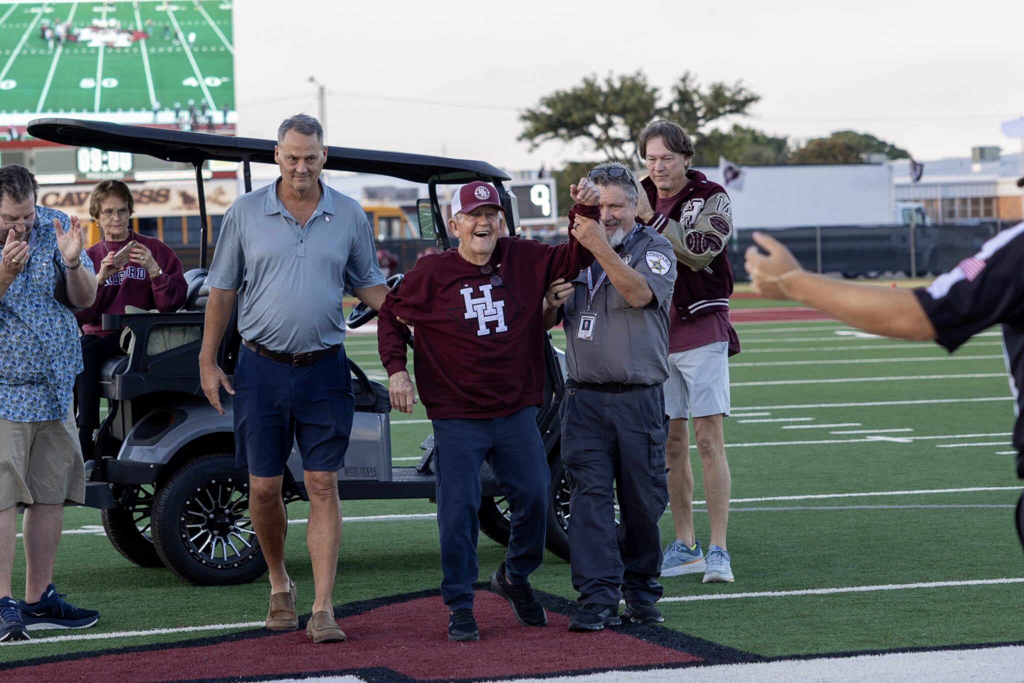 FW9A8451 | Press Pass Sports Group of adults on a football field celebrating near a parked golf cart, with a large scoreboard in the background.
