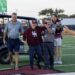 Group of adults on a football field celebrating near a parked golf cart, with a large scoreboard in the background.