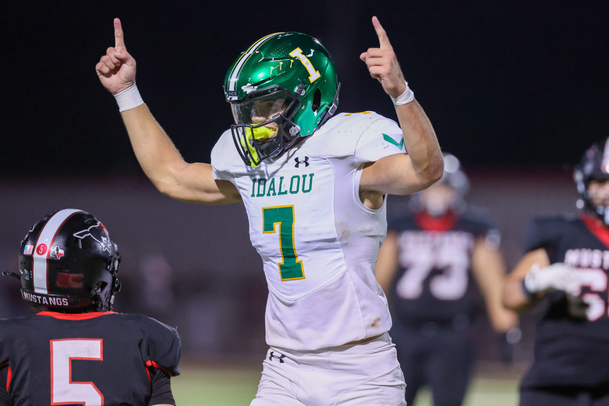 Seabolt Chase-19135 | Press Pass Sports Football player in white Idalou jersey raises both arms in celebration on the field at night, green helmet visible.