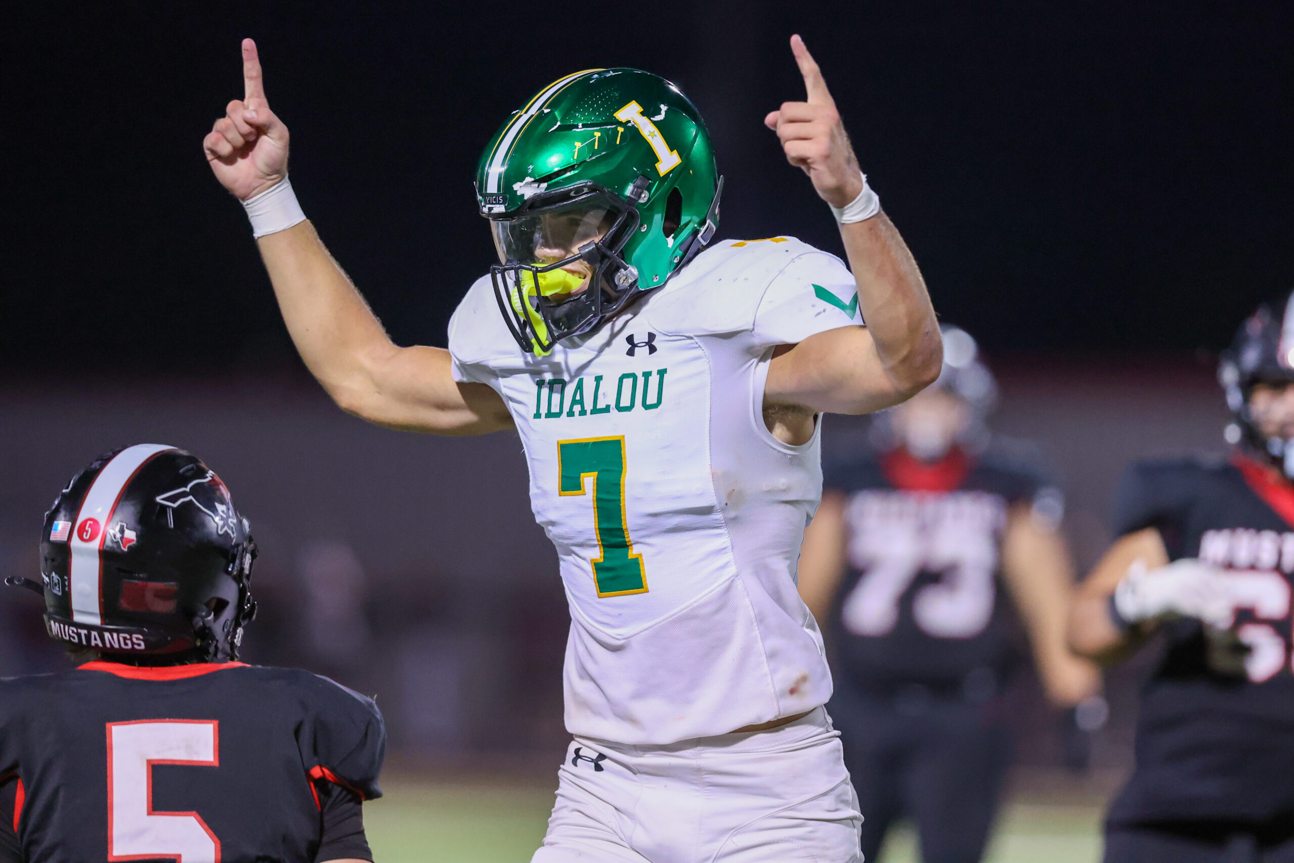 Seabolt Chase-19135 | Press Pass Sports Football player in white Idalou jersey raises both arms in celebration on the field at night, green helmet visible.