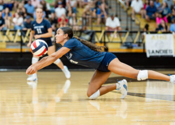 IMG_7725 | Press Pass Sports Female volleyball player in blue dives to save a ball on a polished gym floor with spectators in the stands behind her.