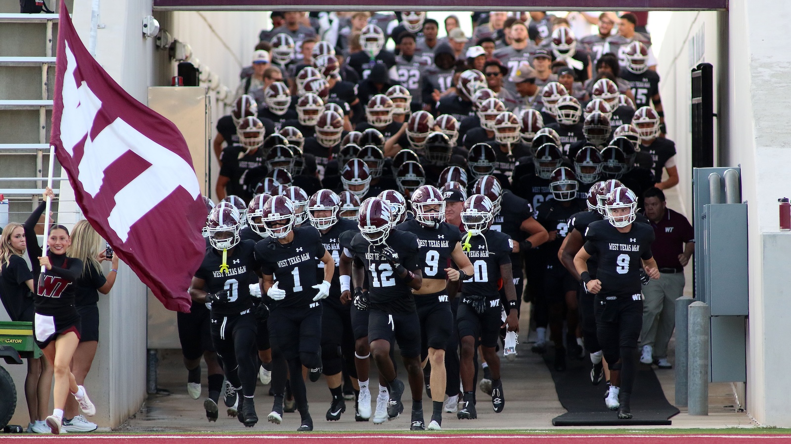 West Texas A&M football team runs onto the field from a tunnel, a large maroon flag waving on the left.