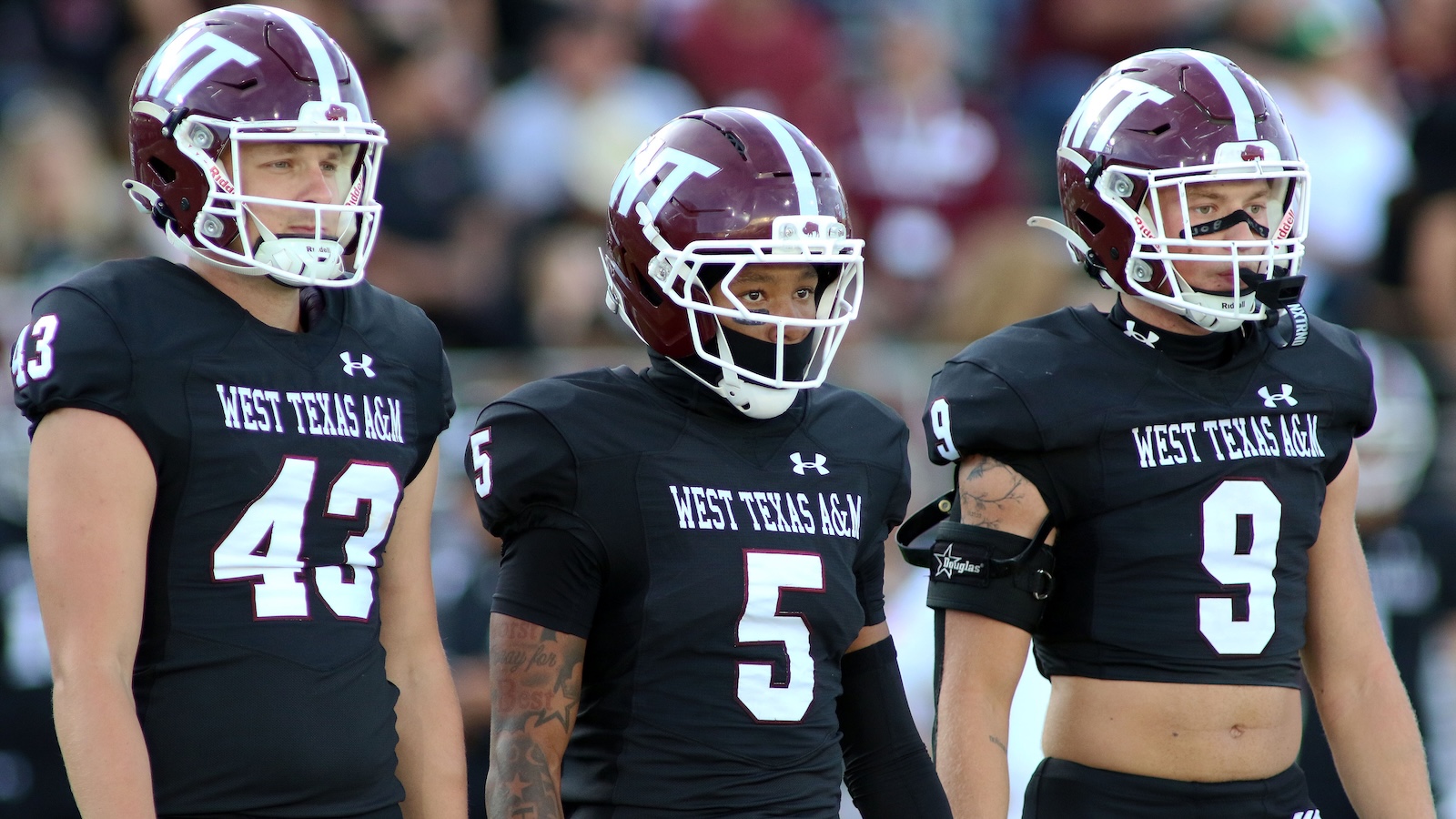 Three West Texas A&M football players stand together in black uniforms with maroon helmets on the field, numbers 43, 5, and 9 visible.