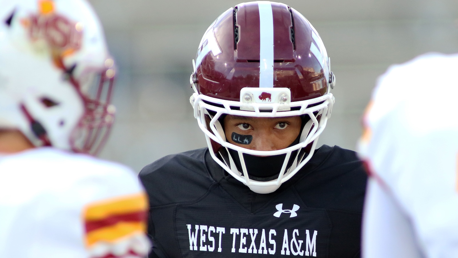 West Texas A&M football player in burgundy helmet with white stripe, black jersey, staring ahead between teammates.