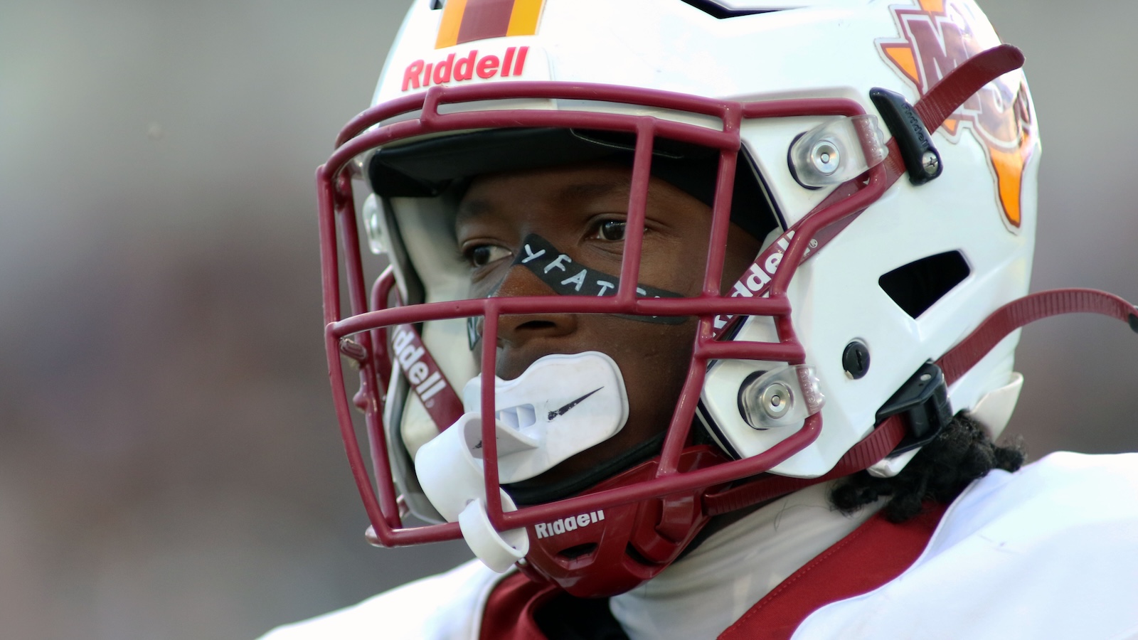 Close-up of a football player's face behind a maroon faceguard on a white helmet during a game.