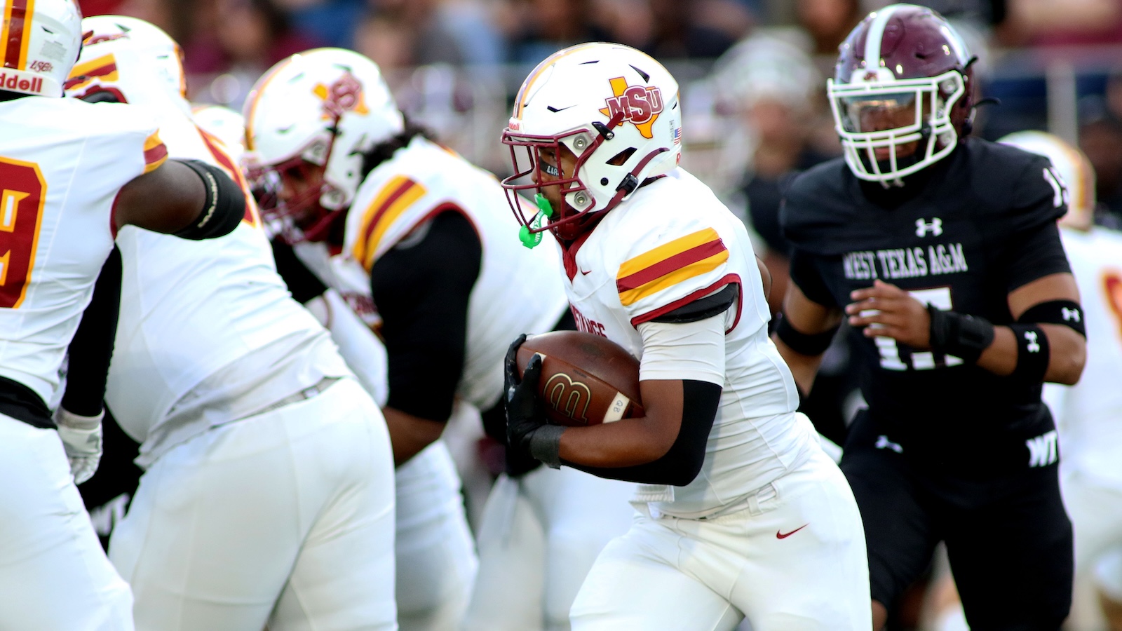 MSU football player in white and gold jersey runs with the ball while teammates block nearby defenders in white and a Black jersey closes in on the right flank.
