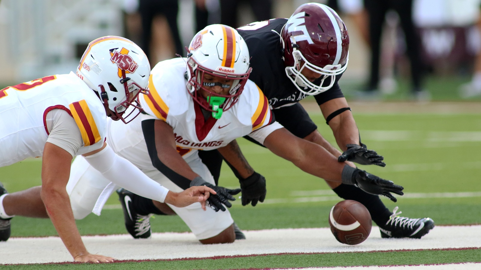 Three football players in white and maroon uniforms dive toward a loose football on the turf.