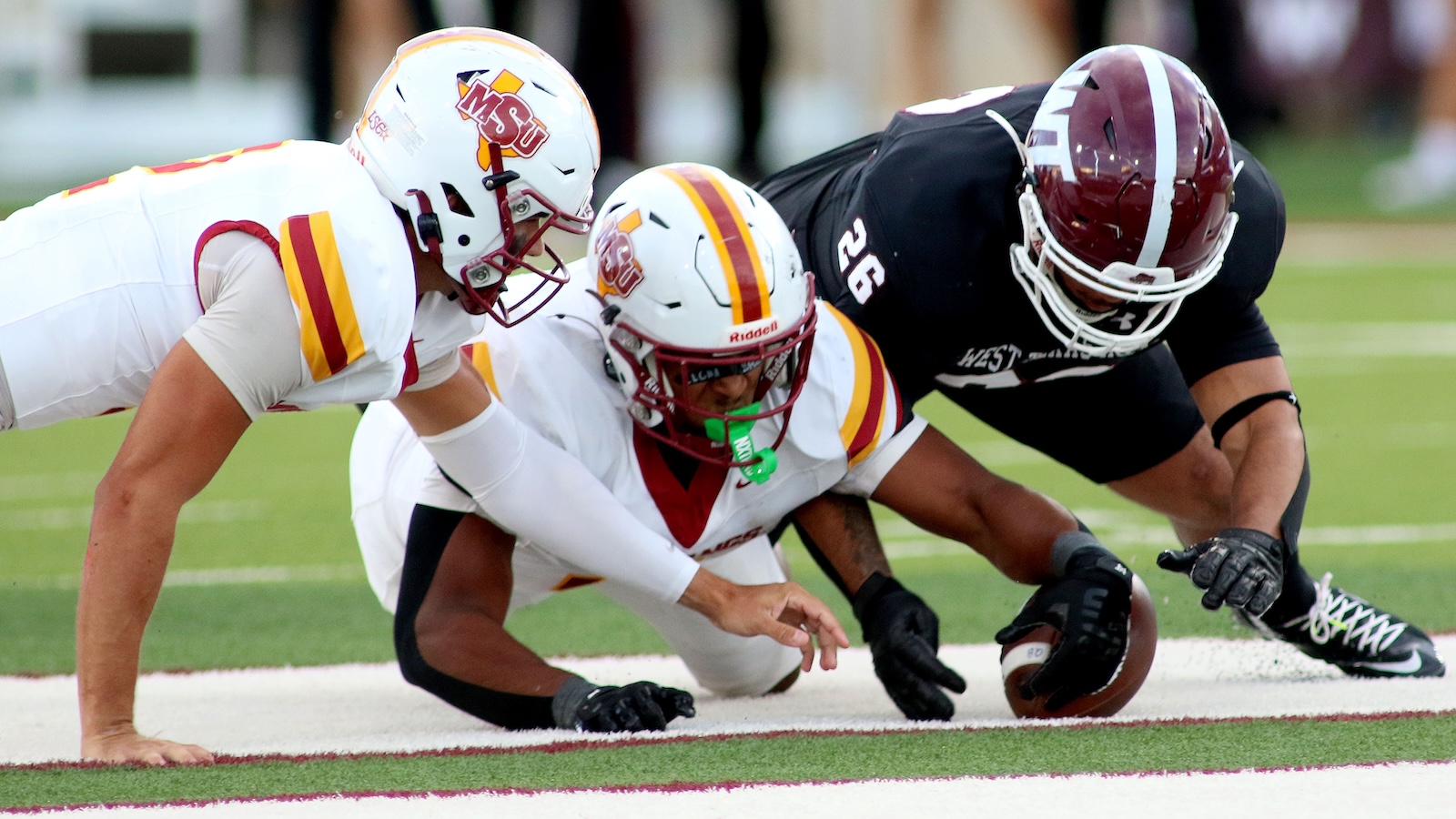 Three football players dive for the ball at the line; two wear white uniforms with MTSU logos and a black-uniform opponent nearby.