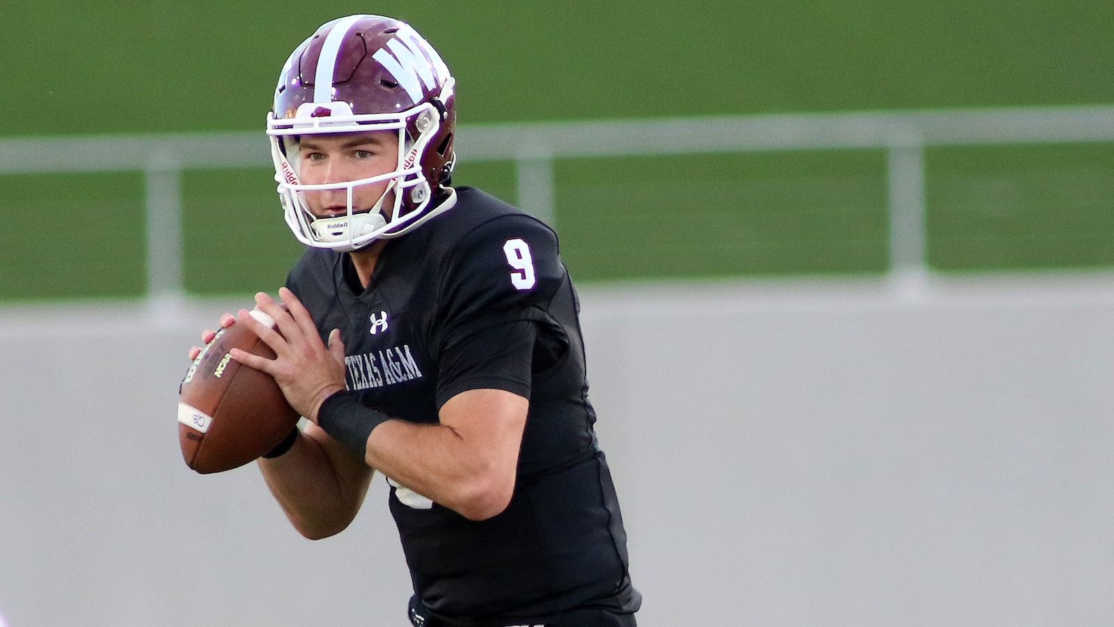 Male football quarterback in black jersey with number 9, gripping a football and looking downfield on a field