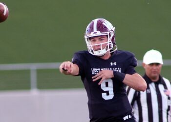 Quarterback in black uniform throws a football during a game, with a referee in stripes in the background.
